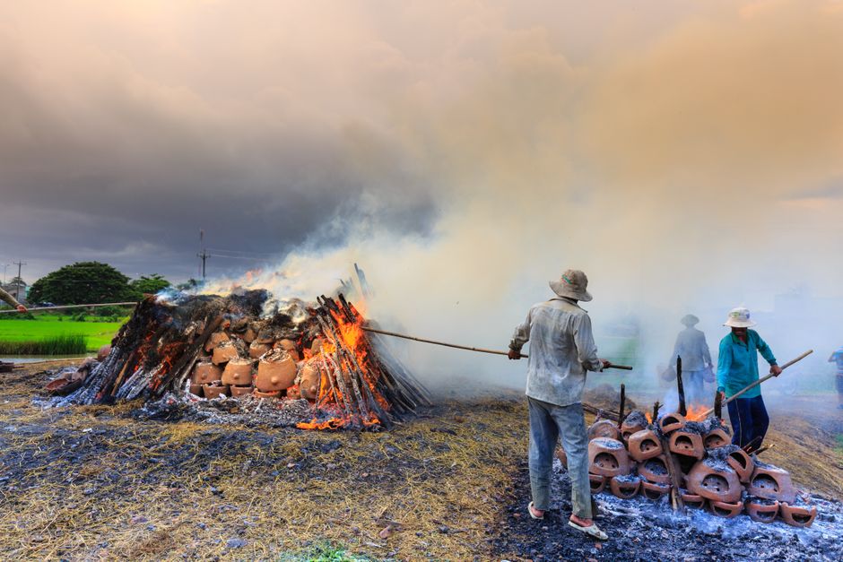 Artisans oversee the firing of clay pots with burning wood in a smoky outdoor setting during sunset.