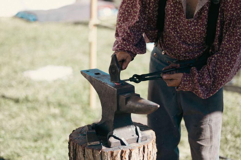 A blacksmith in period clothing is forging metal on an anvil outdoors with tongs and hammer.